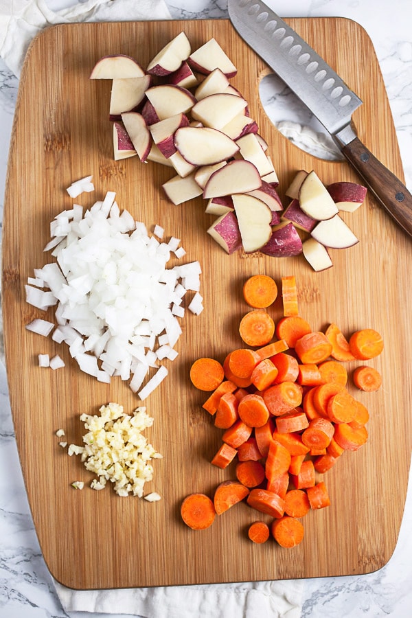 Minced garlic and onions, chopped red potatoes and carrots on wooden cutting board with knife.