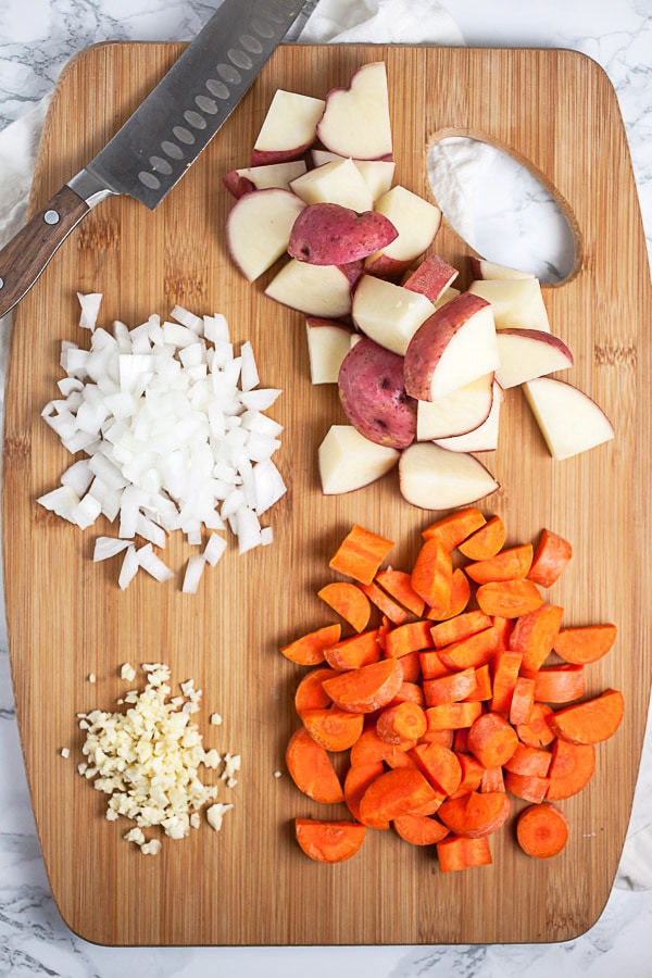 Minced garlic and onions, chopped red potatoes and carrots on wooden cutting board with knife.