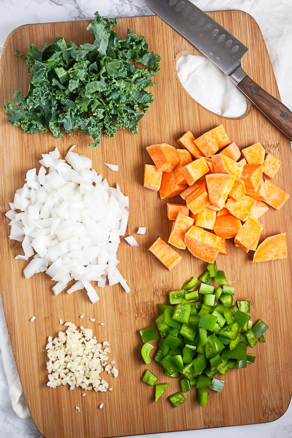 Minced garlic, onions, green bell peppers, kale, and sweet potatoes on wooden cutting board with knife.