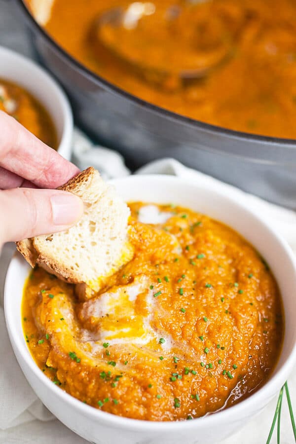 Piece of bread dipped into small white bowl of pureed carrot soup.