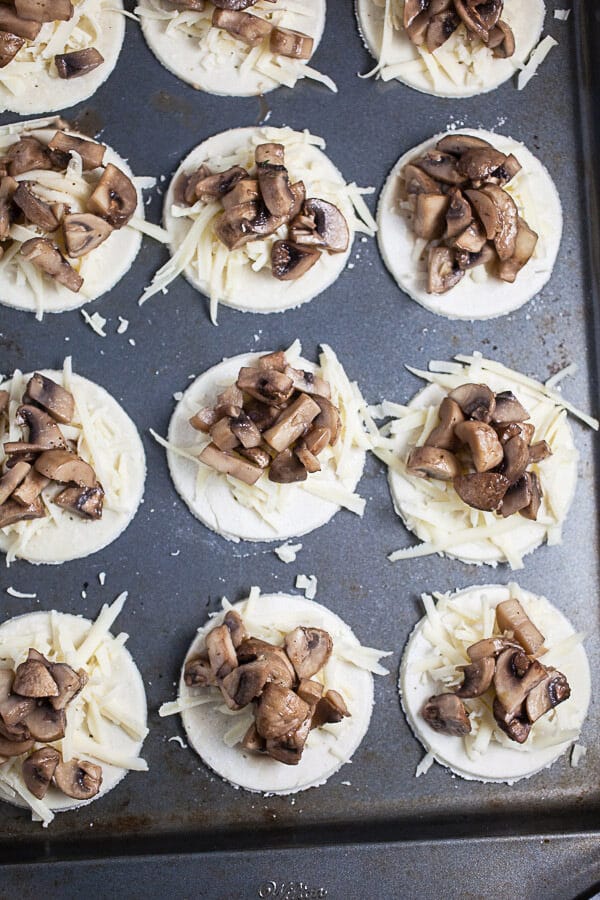 Unbaked tartlets with mushrooms and shredded Gruyere cheese on metal baking sheet.