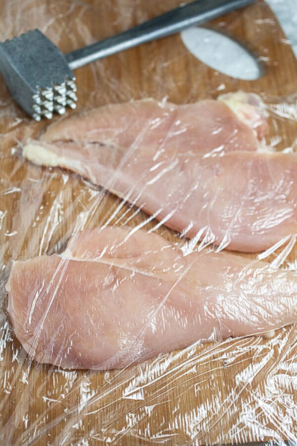 Raw chicken with plastic wrap and meat mallet on wooden cutting board.