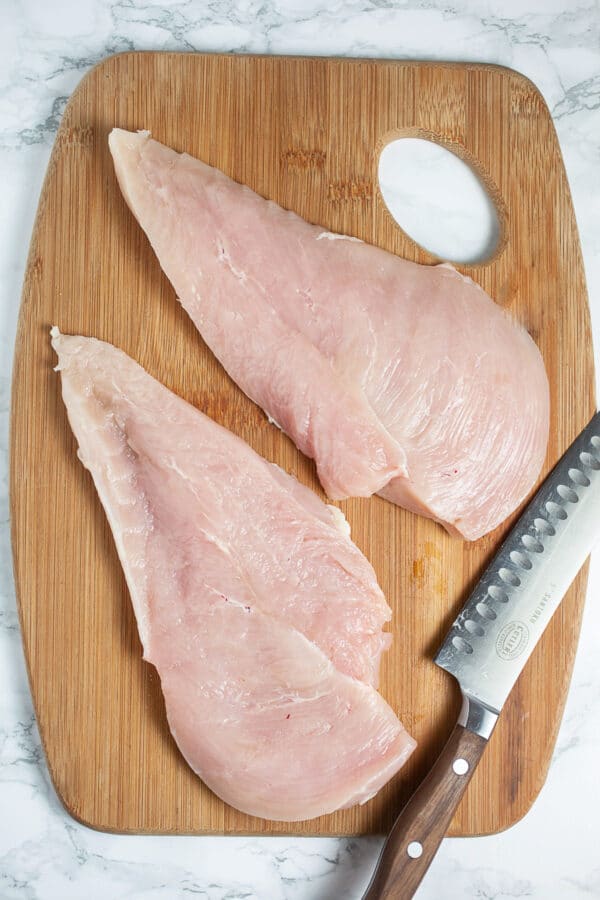 Raw chicken breast cut in half on wooden cutting board with knife.