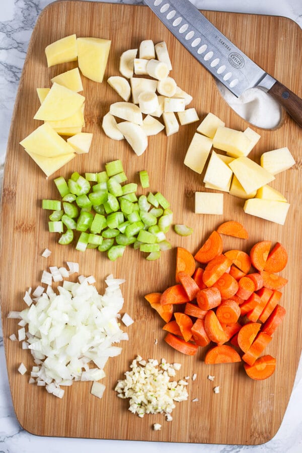 Minced garlic, onions, celery, rutabaga, parsnips, potatoes, and carrots on wooden cutting board with knife.