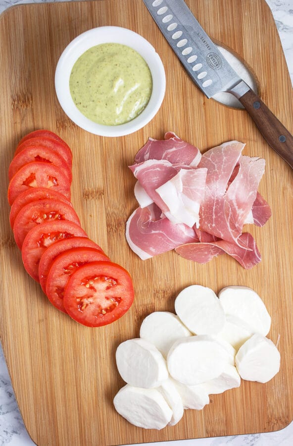 Sliced tomatoes, fresh mozzarella, prosciutto, and basil pesto mayo on wooden cutting board.