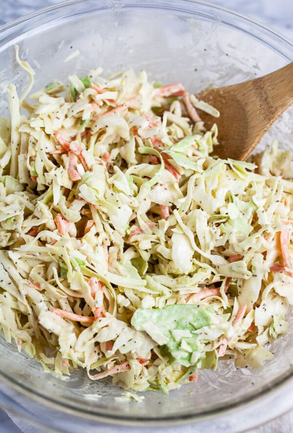 Cabbage coleslaw mixed with dressing in large glass bowl with wooden spoon.