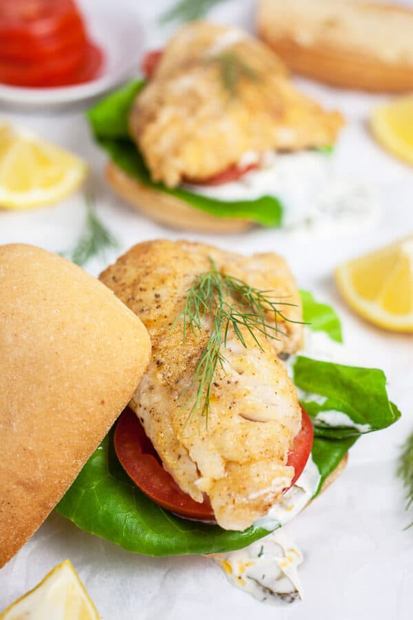 Walleye sandwiches on buns with tartar sauce, lettuce, tomatoes, fresh dill, and lemon wedges.