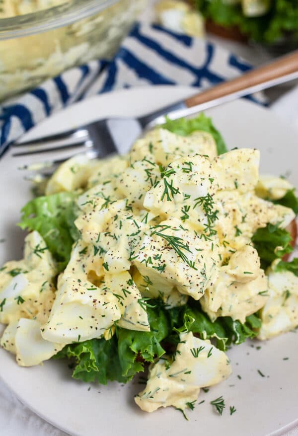 Egg salad on bread and lettuce garnished with fresh dill on small white plate with fork.