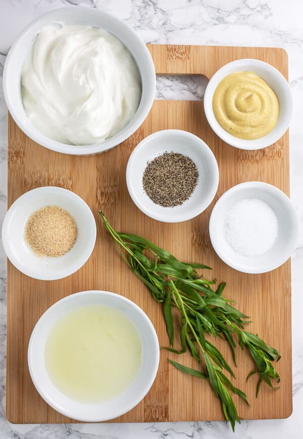 Lemon juice, garlic powder, salt, pepper, sour cream, Dijon mustard, and fresh tarragon on wooden cutting board.