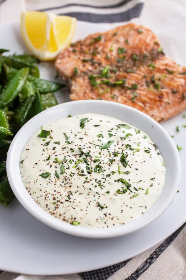 Small bowl of tarragon sauce on white plate with salmon, peas, and lemon wedge.