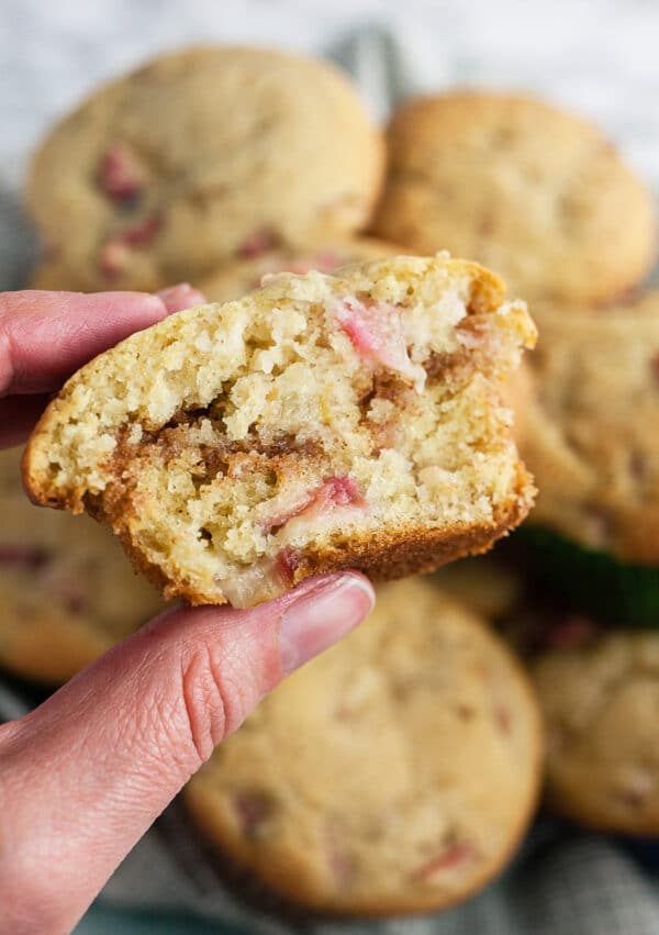 Fingers holding streusel rhubarb muffin cut in half in front of bowl of muffins.