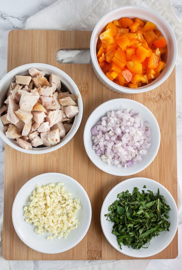 Garlic, chicken, orange bell peppers, shallots, and basil in small white bowls on wooden cutting board.