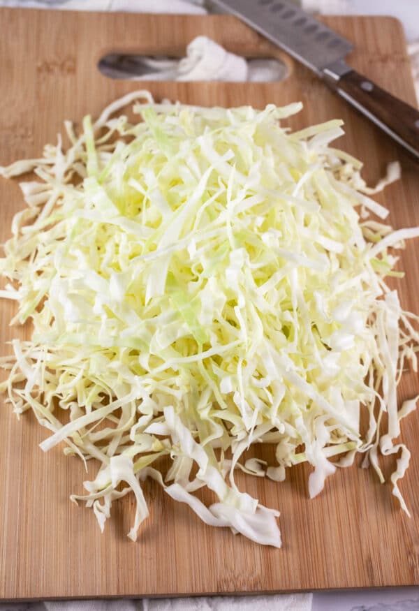 Shredded green cabbage on wooden cutting board with knife.