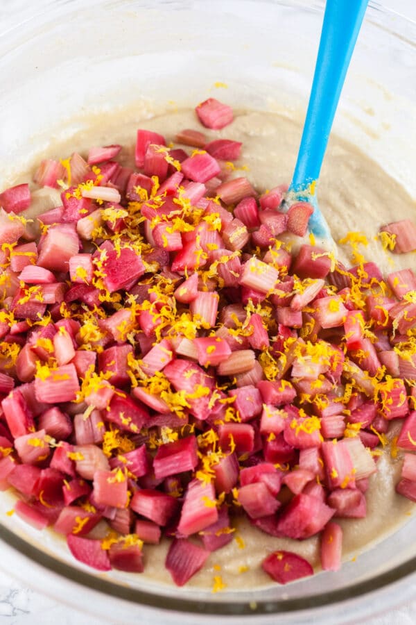 Cake batter with sliced rhubarb, orange, and lemon zest in large glass bowl with blue spatula.