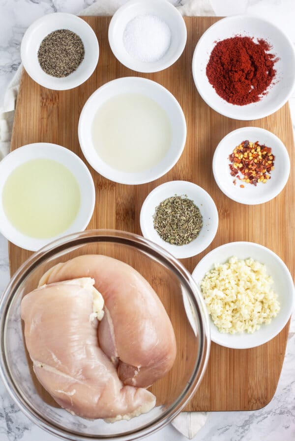 Chicken breasts, minced garlic, oil, vinegar, and spices in bowls on wooden cutting board.