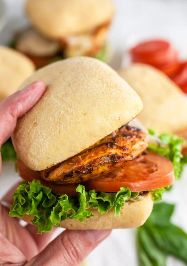 Hand holding grilled chicken breasts sandwich on bun with lettuce and tomato.