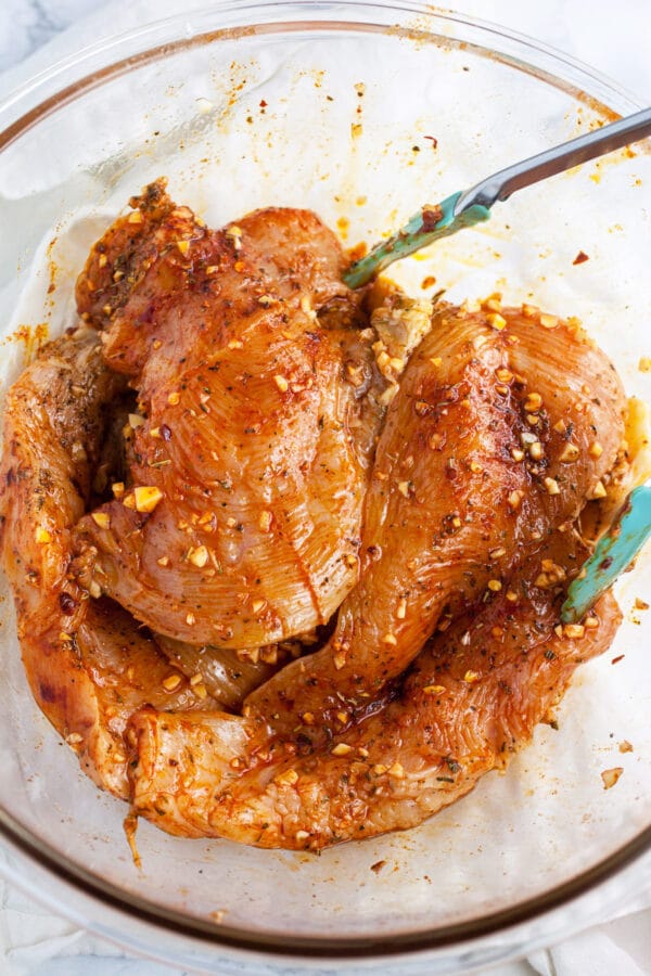 Chicken breasts marinated in oil, garlic, and spices in large glass bowl.