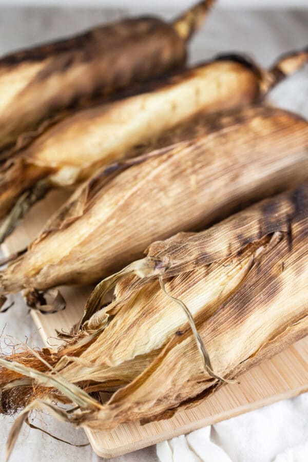 Grilled corn cobs in husks on wooden cutting board.