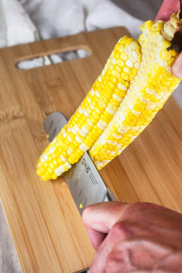 Grilled corn being cut from cob with a knife on wooden cutting board.