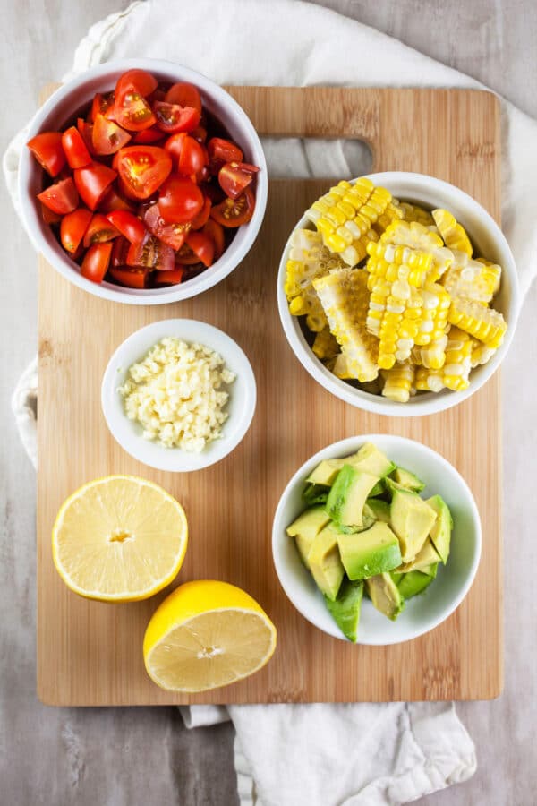 Grilled corn, tomatoes, garlic, avocadoes, and lemon in bowls on wooden cutting board.