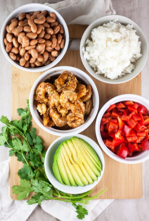 Sautéed shrimp, rice, pinto beans, red bell peppers, avocado, and cilantro in bowls on wooden cutting board.