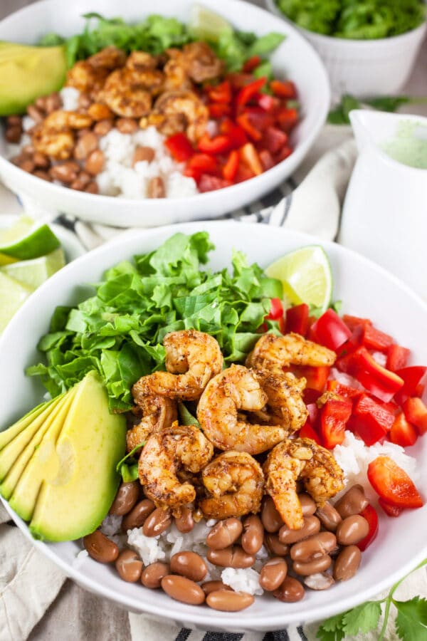 Sautéed shrimp, red bell peppers, lettuce, avocado, pinto beans, and lime in white bowls.