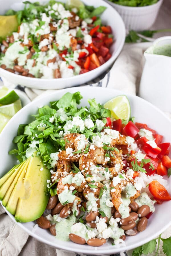 These Mexican Shrimp Rice Bowls are the perfect quick and easy healthy dinner recipe! Rice forms the base of this meal and it's topped with spicy Mexican shrimp, black beans, avocado, and a simple creamy cilantro lime dressing. This delicious dressing is made with sour cream. I love a hearty and flavorful rice bowl! #shrimp #ricebowl #Mexican #cilantro #lime Mexican shrimp rice bowls with red peppers, lettuce, avocado, and cilantro lime cream sauce with queso fresco cheese.