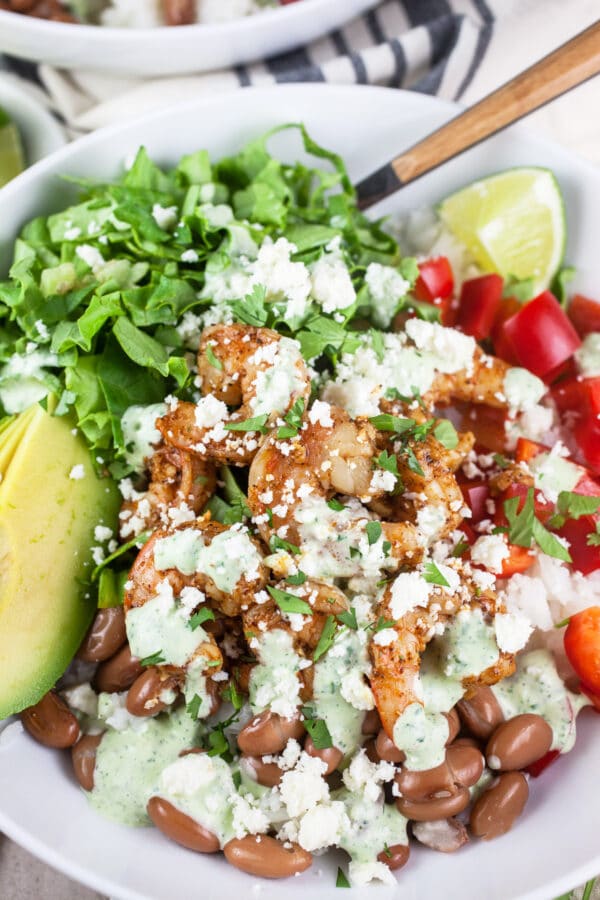 Cooked shrimp, pinto beans, red bell peppers, lettuce, avocado, and lime topped with creamy dressing and queso fresco cheese in white bowl.