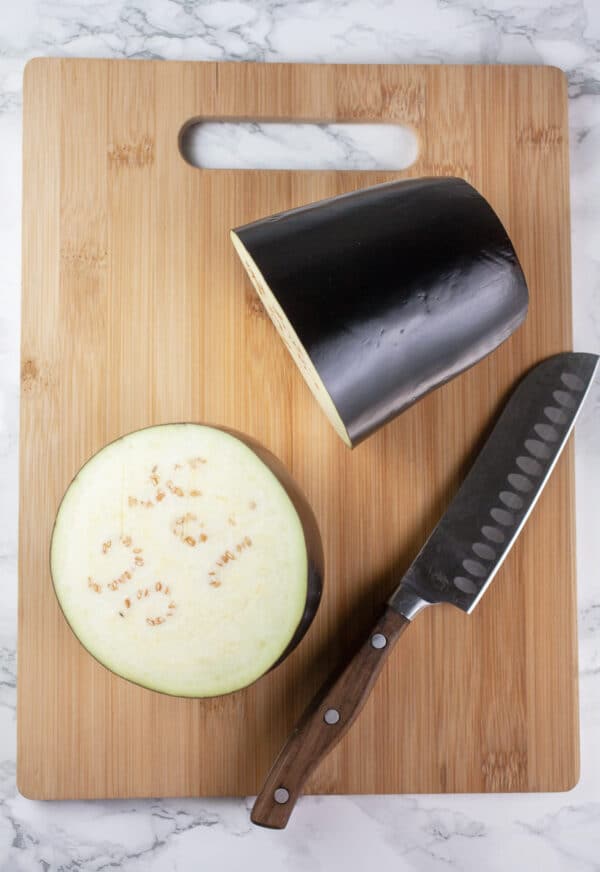 Eggplant cut in half on wooden cutting board with knife.