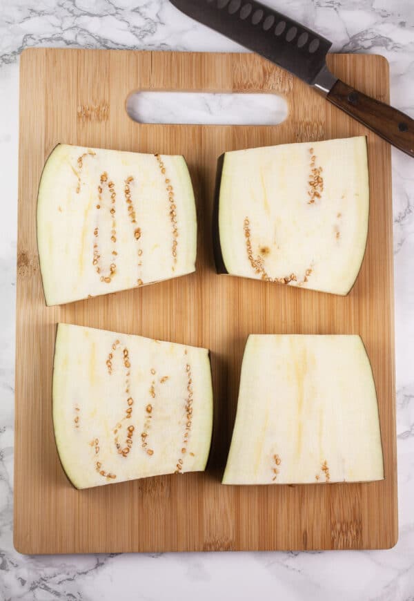 Eggplant halves cut in half on wooden cutting board with knife.