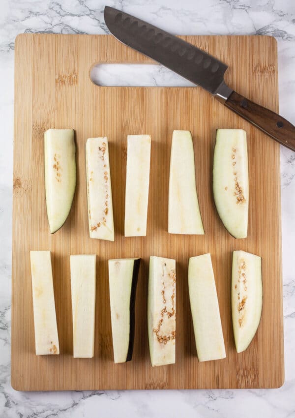 Eggplant cut into fries on wooden cutting board with knife.