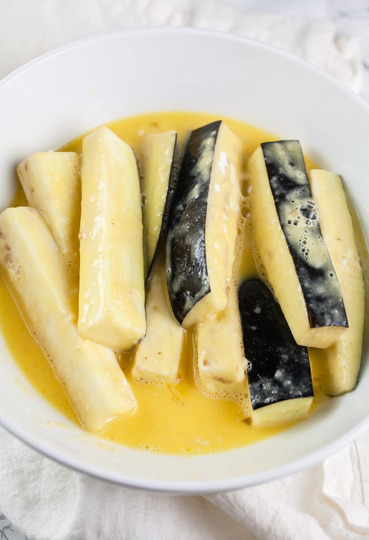 Eggplant fries coated in egg mixture in white bowl.
