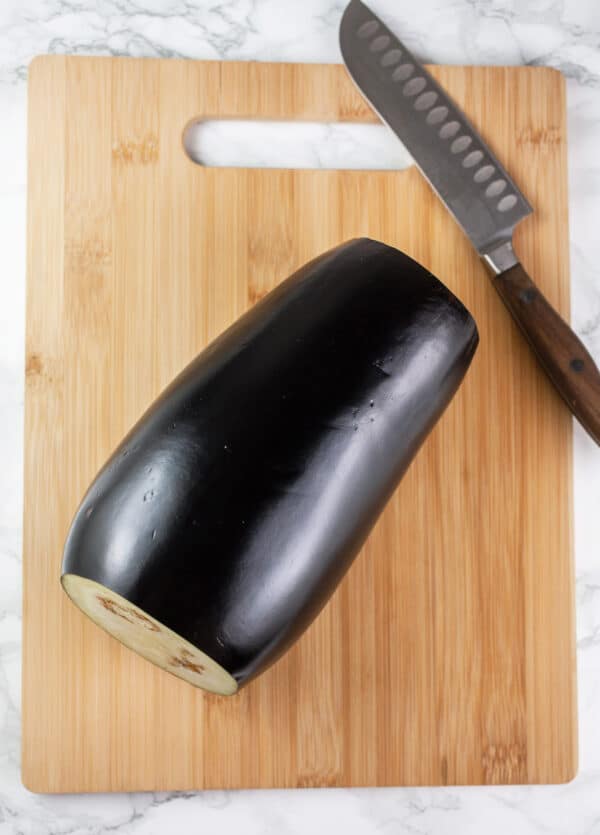 Eggplant with top and bottom removed on wooden cutting board with knife.