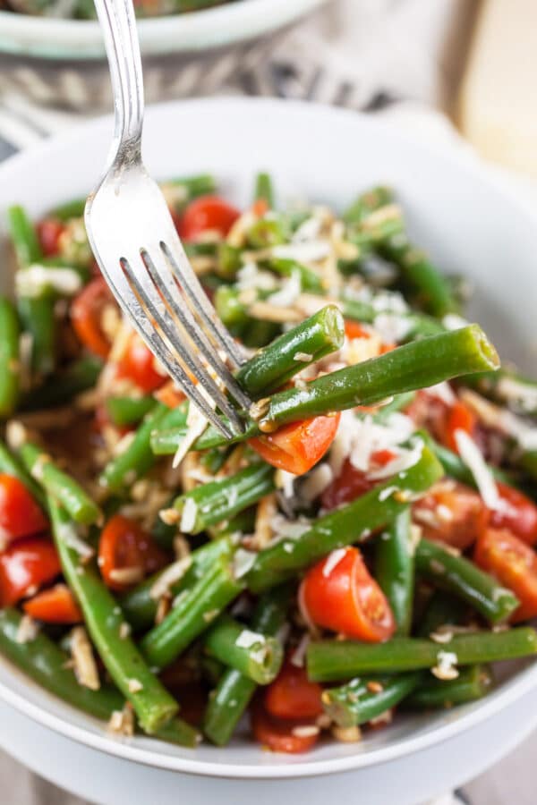 Fork holding green bean tomato salad in front of white bowl.