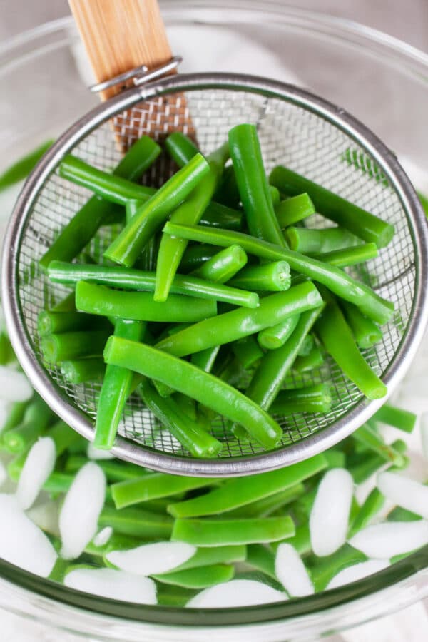 Blanched green beans in strainer lifted from glass bowl of ice water.