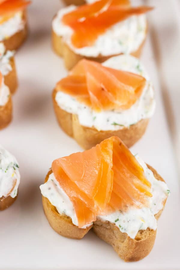 Smoked salmon slices on ricotta topped crostini on white serving platter.