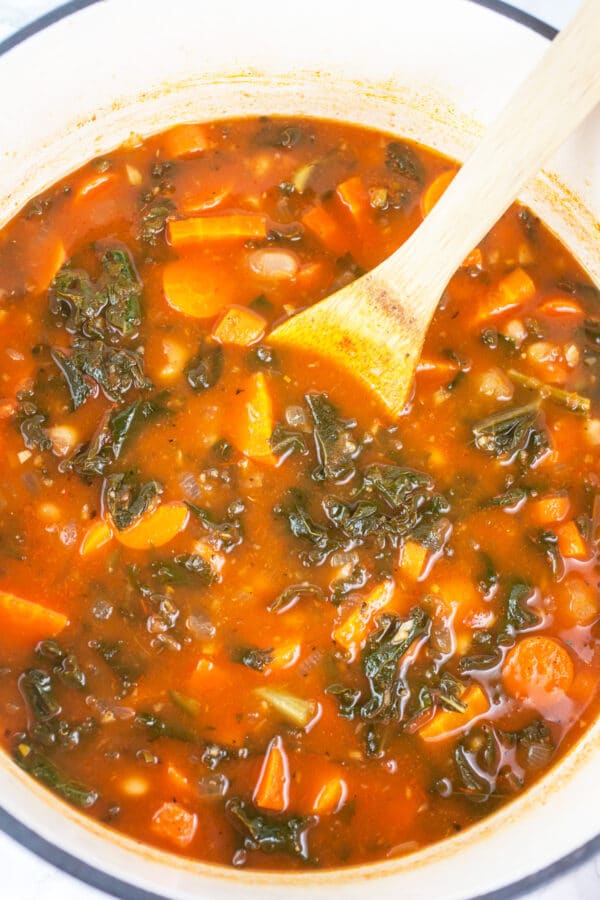 Cooked cannellini bean, kale, and carrot soup in white cast iron Dutch oven.