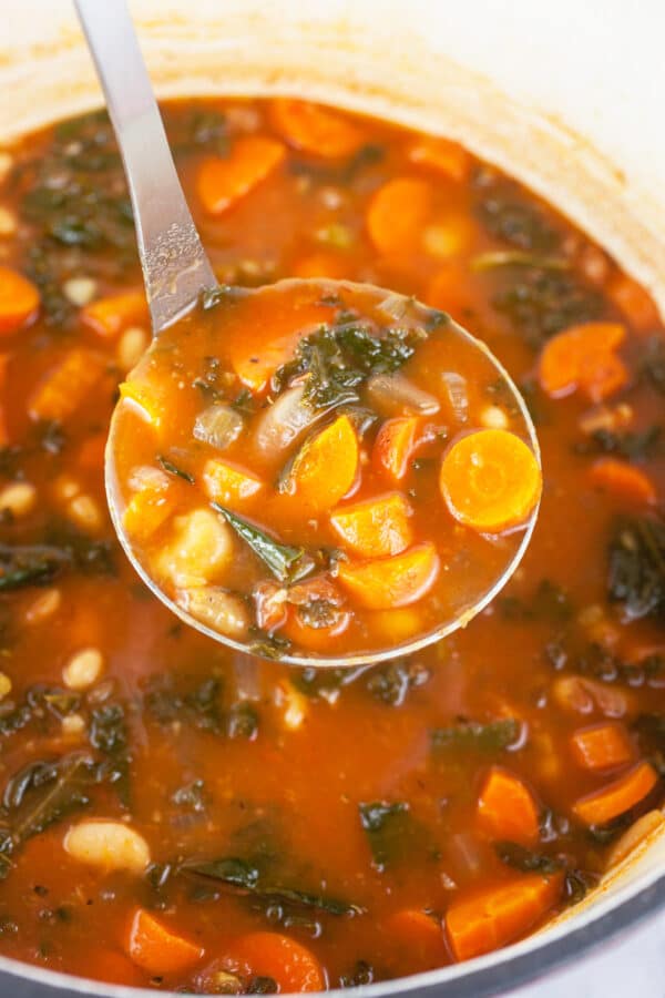 Scoop of white bean, kale, and carrot soup lifted from pot with soup ladle.