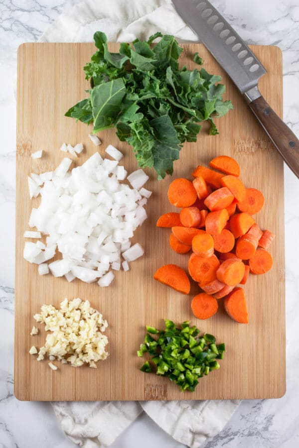 Minced garlic, onions, jalapeno pepper, carrots, and kale on wooden cutting board with knife.