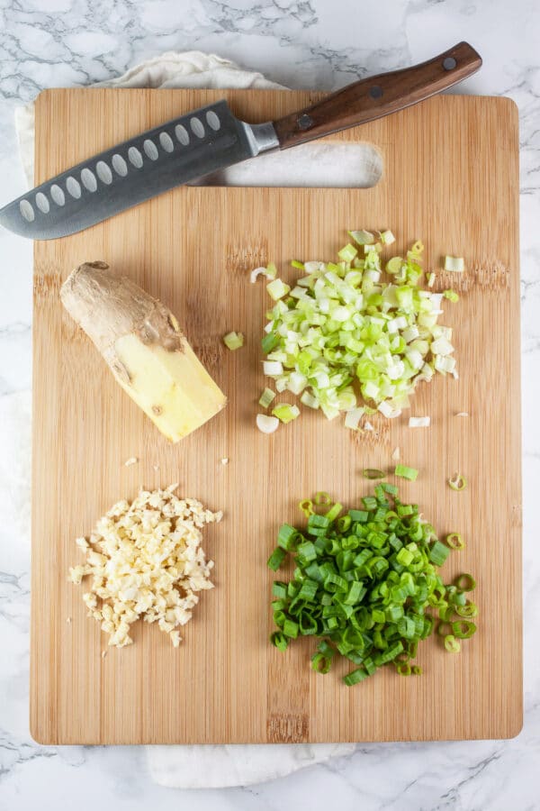 Minced garlic, green onion tops and bottoms, and peeled ginger root on wooden cutting board with knife.