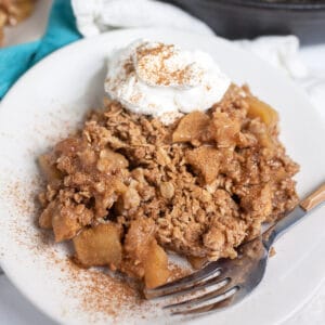 Cast iron apple crisp with whipped cream and cinnamon on small white plate with fork.