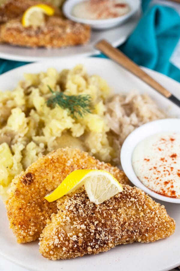 Breaded, fried kohlrabi schnitzel with mashed potatoes, sauerkraut, and dipping sauce on white plate.