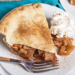 Piece of gluten free apple pie with whipped cream on small white plate with fork.