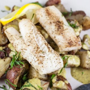 Sheet pan cod and potatoes with pesto sauce and lemon on white plate.