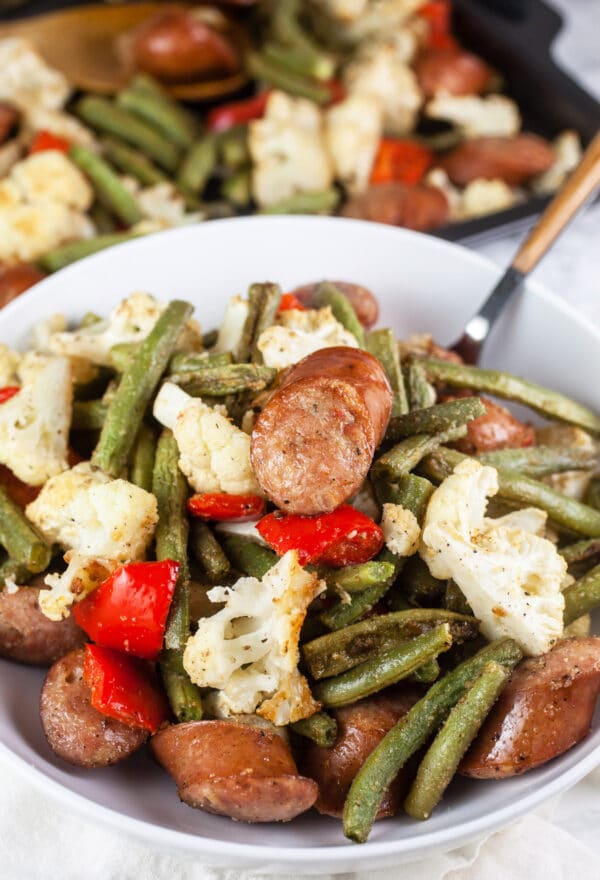 Sheet pan chicken sausage with green beans, cauliflower, and red peppers in white bowl.