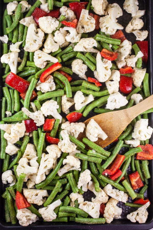 Uncooked green beans, cauliflower, and red bell peppers tossed in olive oil and spices on baking sheet.