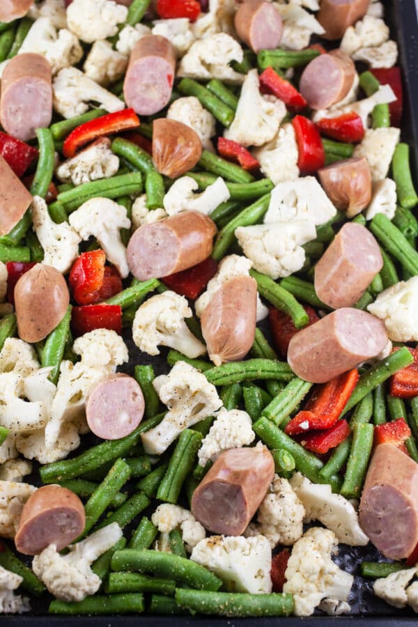 Uncooked chicken sausages, green beans, cauliflower, and red bell peppers on baking sheet.