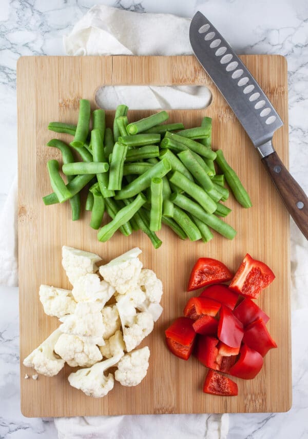 Diced cauliflower, green beans, and red bell peppers on wooden cutting board with knife.