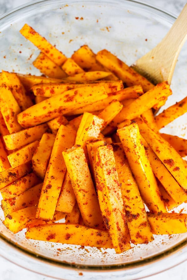 Uncooked squash fries tossed in olive oil and spices in large glass bowl.