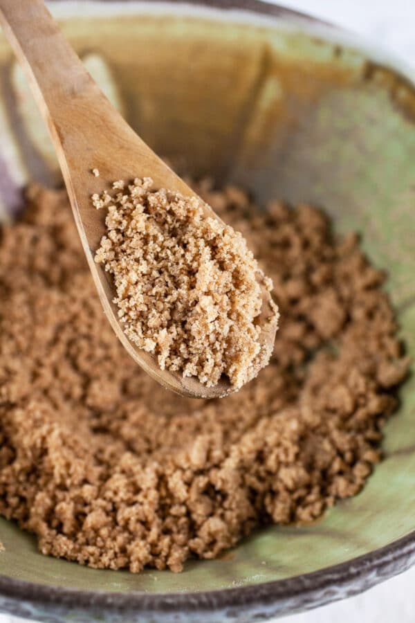 Cinnamon streusel topping in wooden spoon lifted from green ceramic bowl.